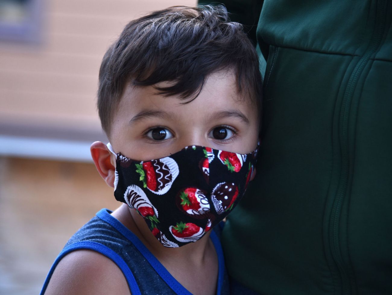 A boy, center, wearing a mask and looking directly into the camera while snuggled against his mother's hip