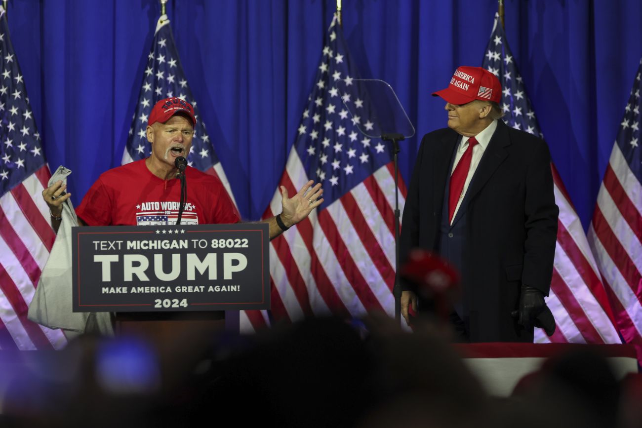 A UAW member speaks at a podium while Donald Trump stands next to him. 