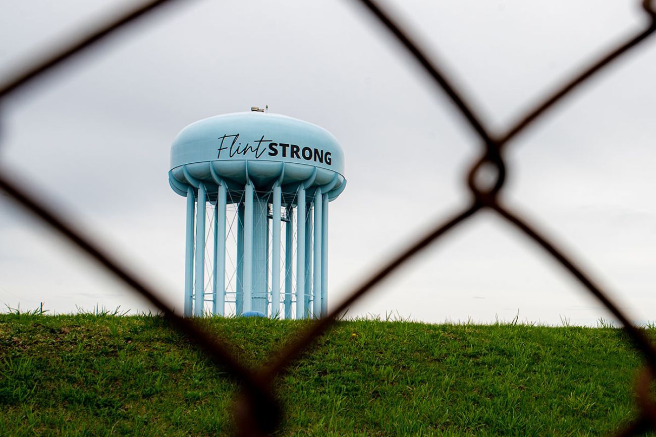 A water tower reading Flint Strong, seen through an iron fence in the foreground