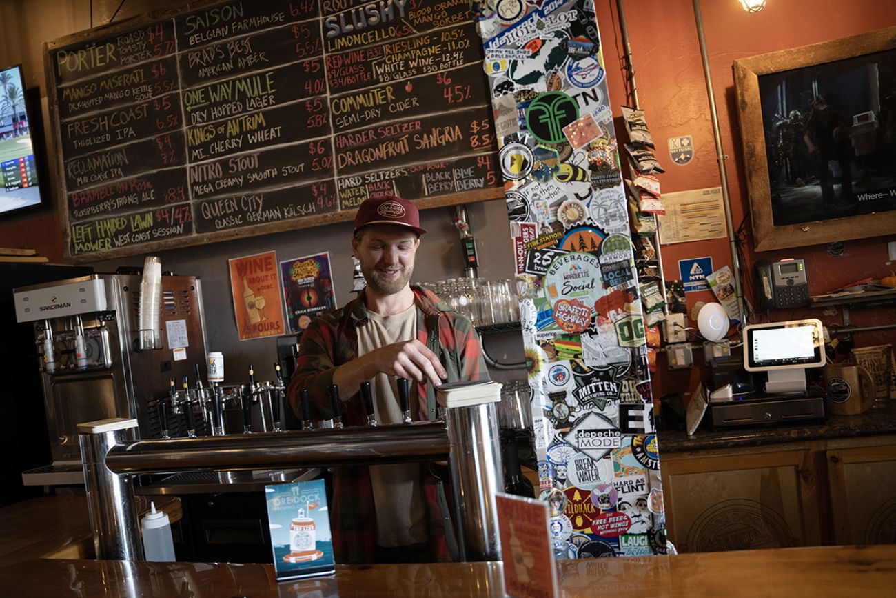 Alec Bush, wearing a baseball hat, pours a beer at Ore Docks Brewing Co. There's a chalkboard behind him of the beers on tap