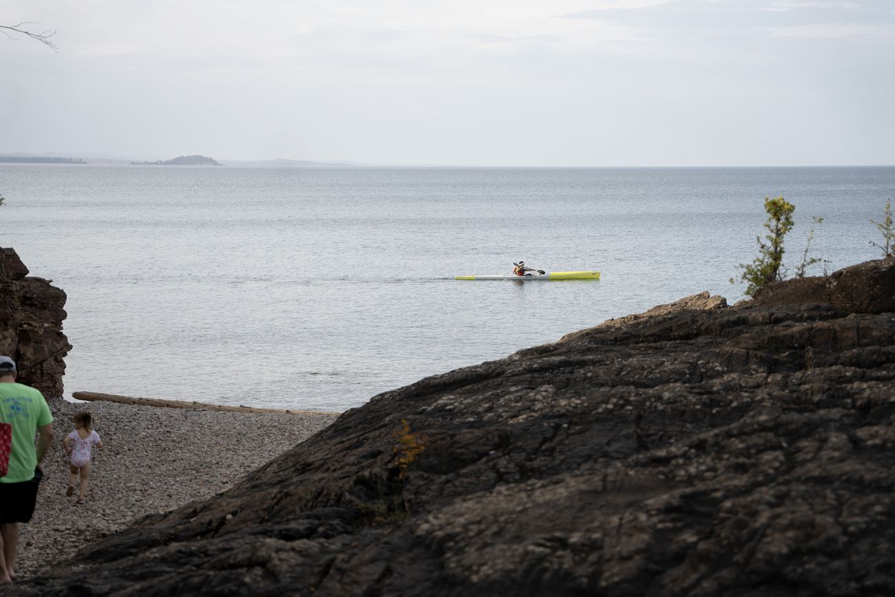 A lone kayaker in a yellow boat past the Black Rocks at Presque Isle Park just north of downtown Marquette. There are a couple of other people walking on the sand