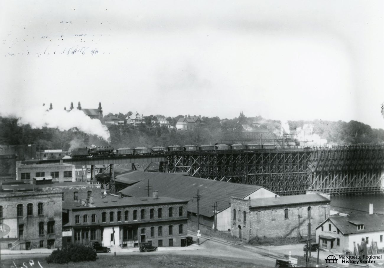 Black and white photo: A train along with a number of train cars delivering ore can be seen atop the train trestle leading to the Ore Dock in the Lower Harbor in Marquette.