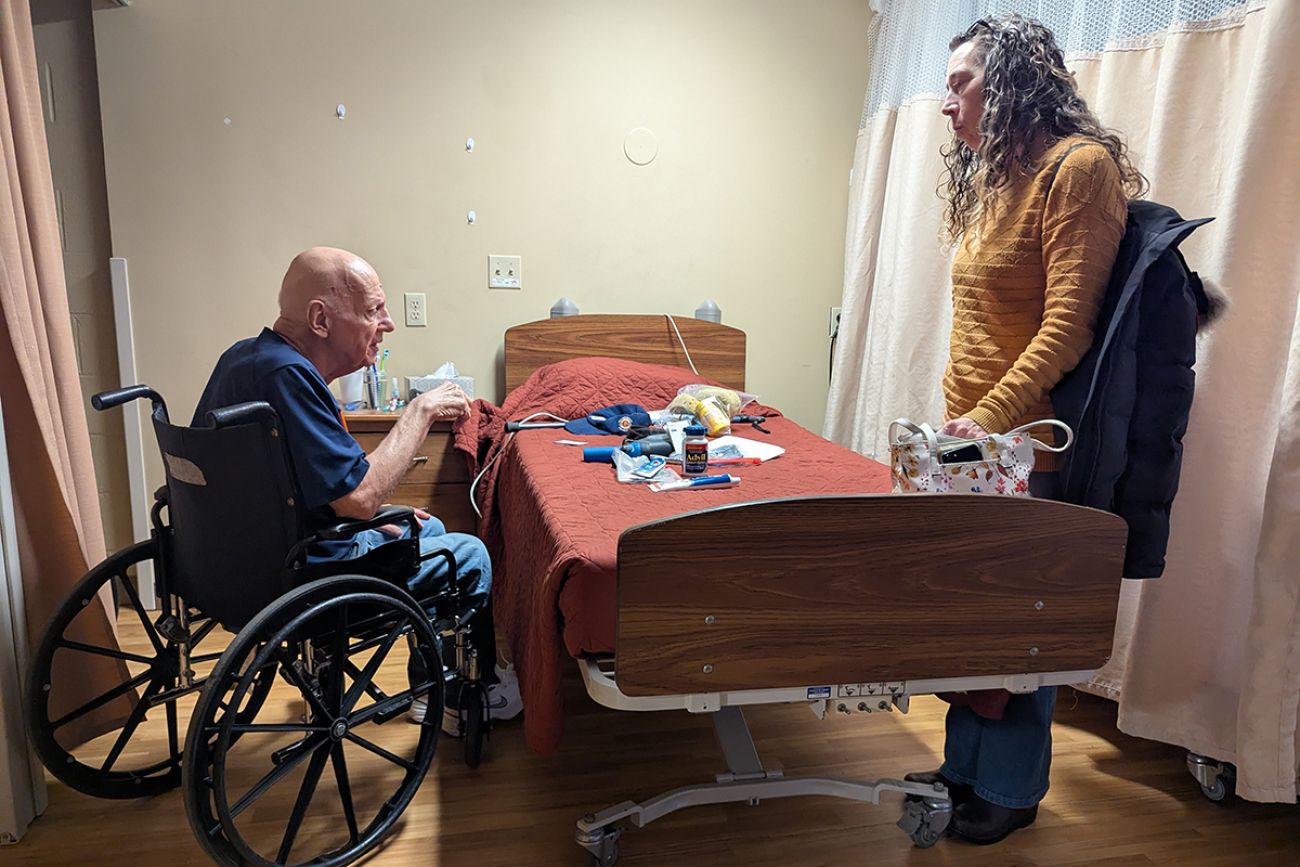Howard Gregor in a wheelchair by a medical bed. A woman is standing on the other side of the bed.