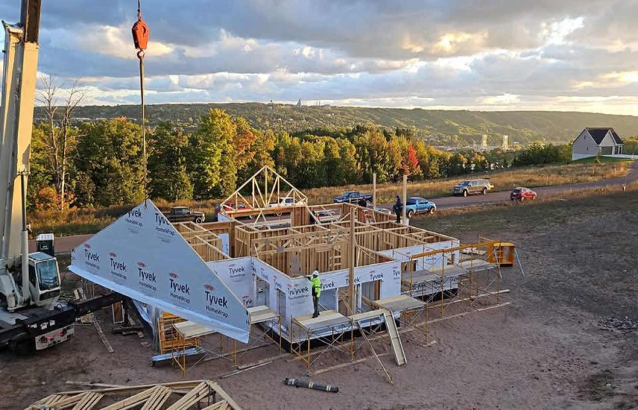 A crane raises a frame to the top of a house that is under construction
