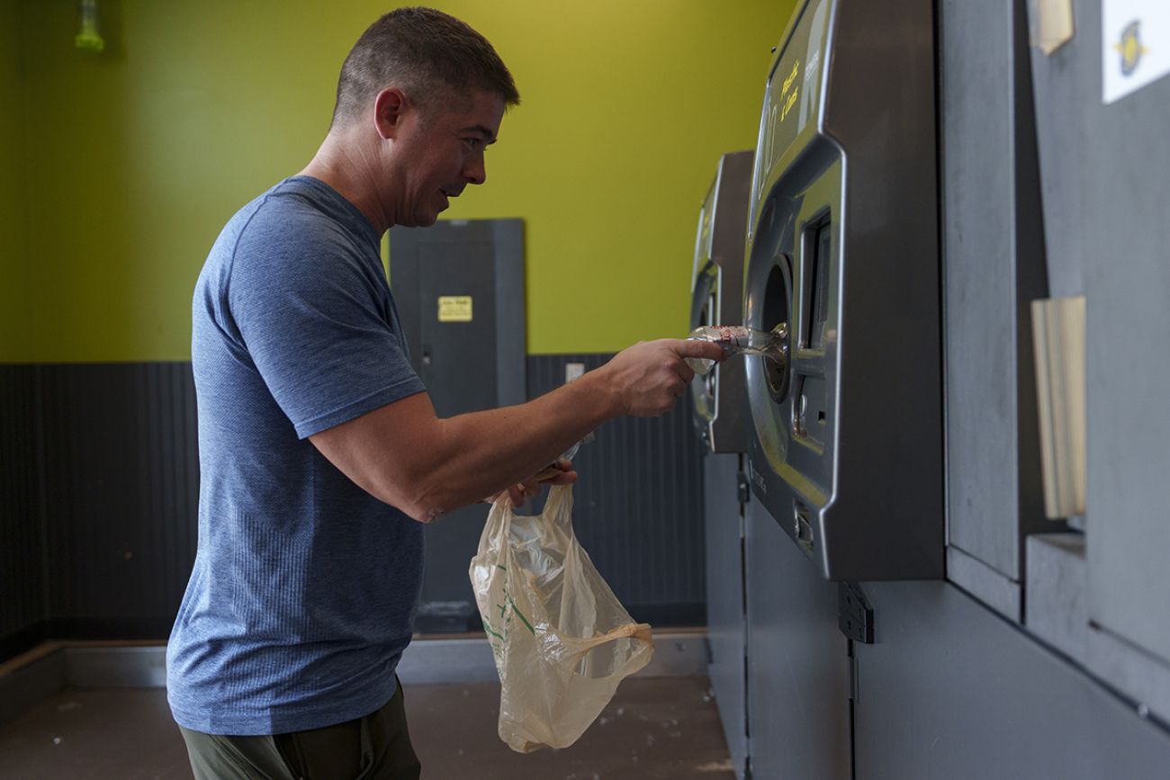 A man in a blue t-shirt places a plastic bottle into a bottle deposit machine.