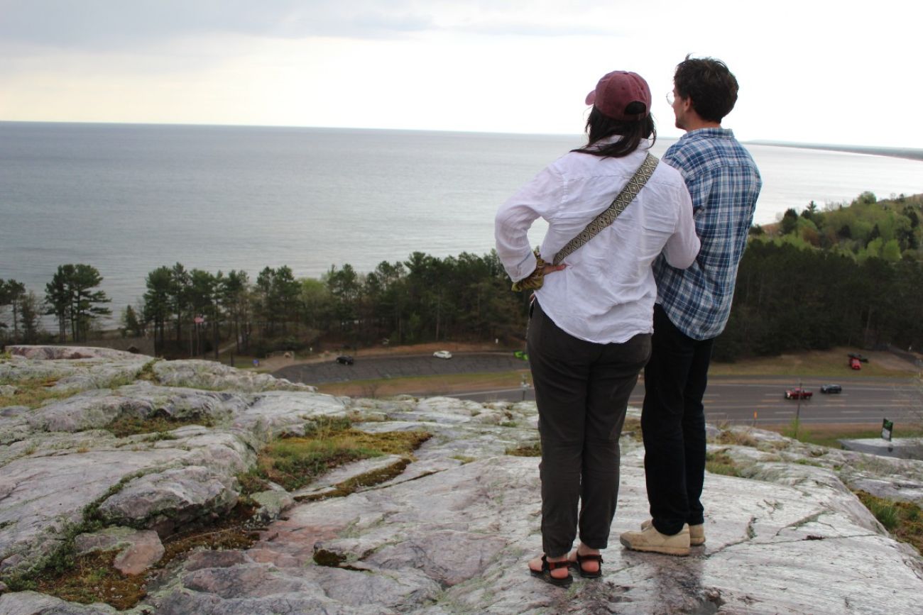 Two people stand at the top of a rock cliff overlooking Lake Superior