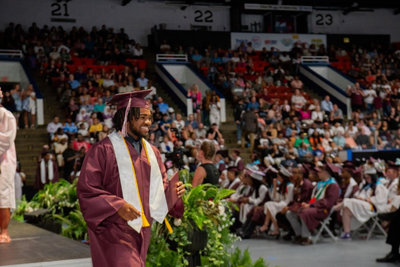 A young man in cap and gown picks up his high school diploma in an auditorium