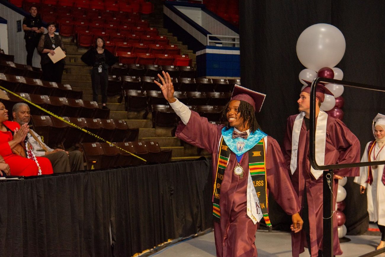 A student in cap and gown waves to the crowd in an auditorium at a graduation ceremony