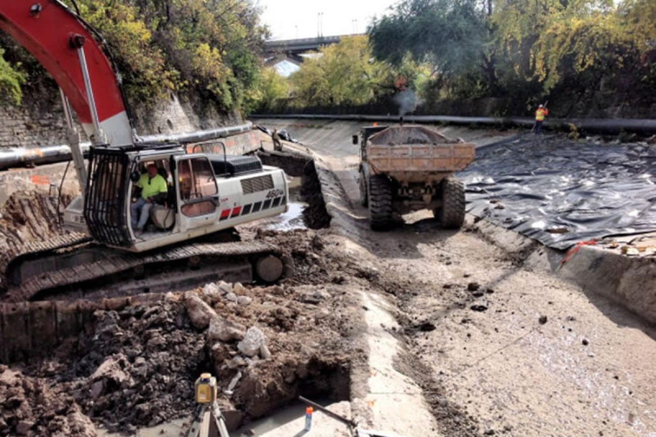 Workers operating heavy construction equipment work in a drained concrete riverbed