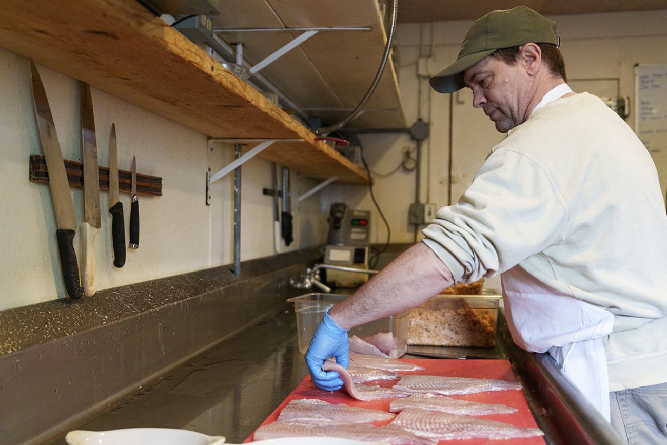 Chris Smith preps whitefish filets on a cutting board in a kitchen. He is wearing a baseball hat and a white chef's coat.