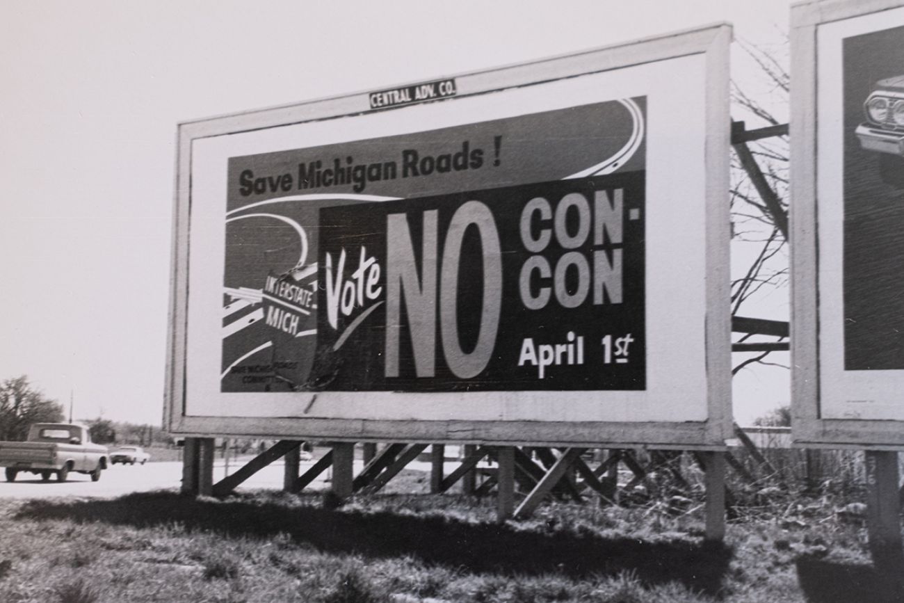 A sign that says "Save Michigan Roads!" It's a black-and-white photo.