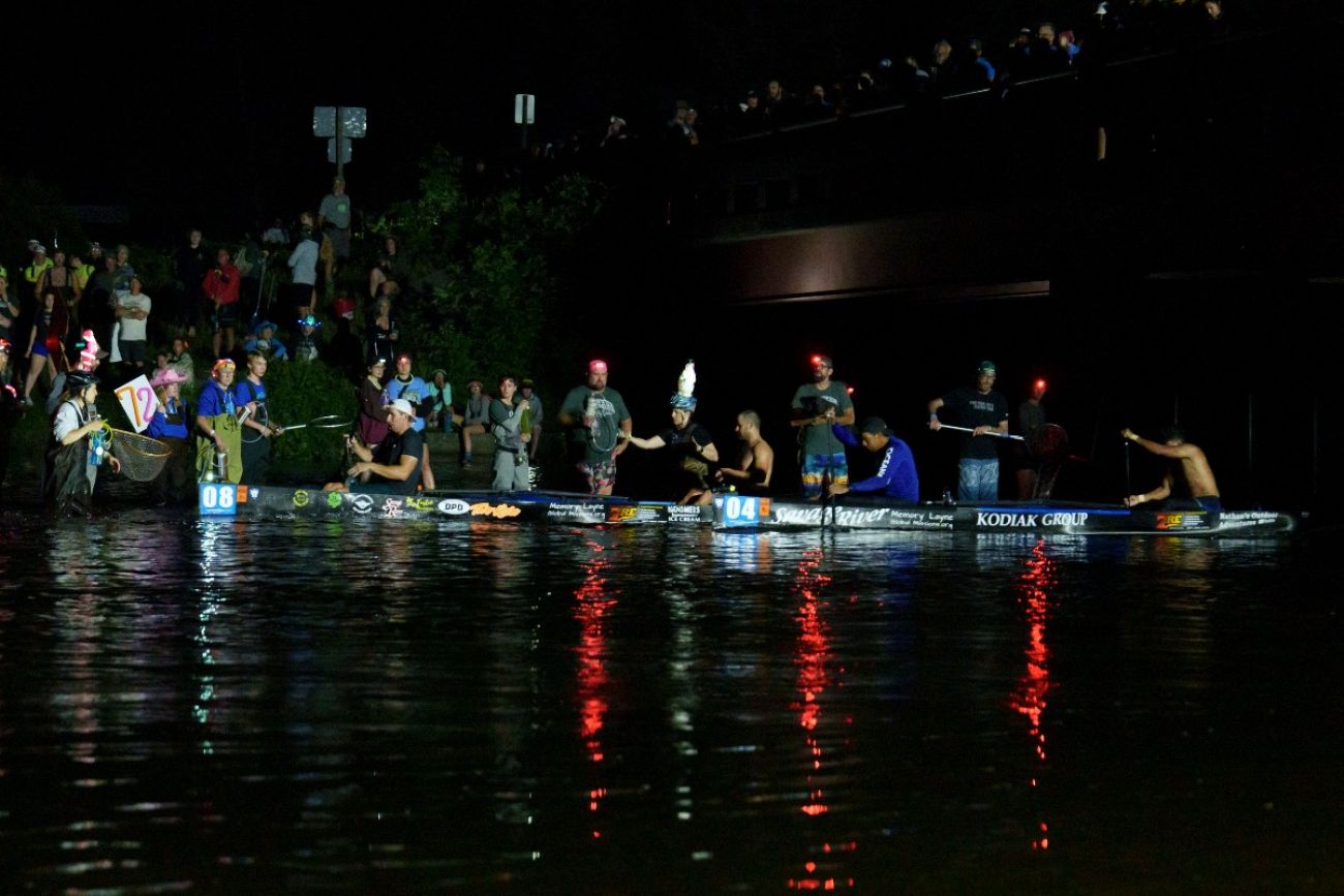 Canoe racers line up along the shore as people on shore pass them food and water