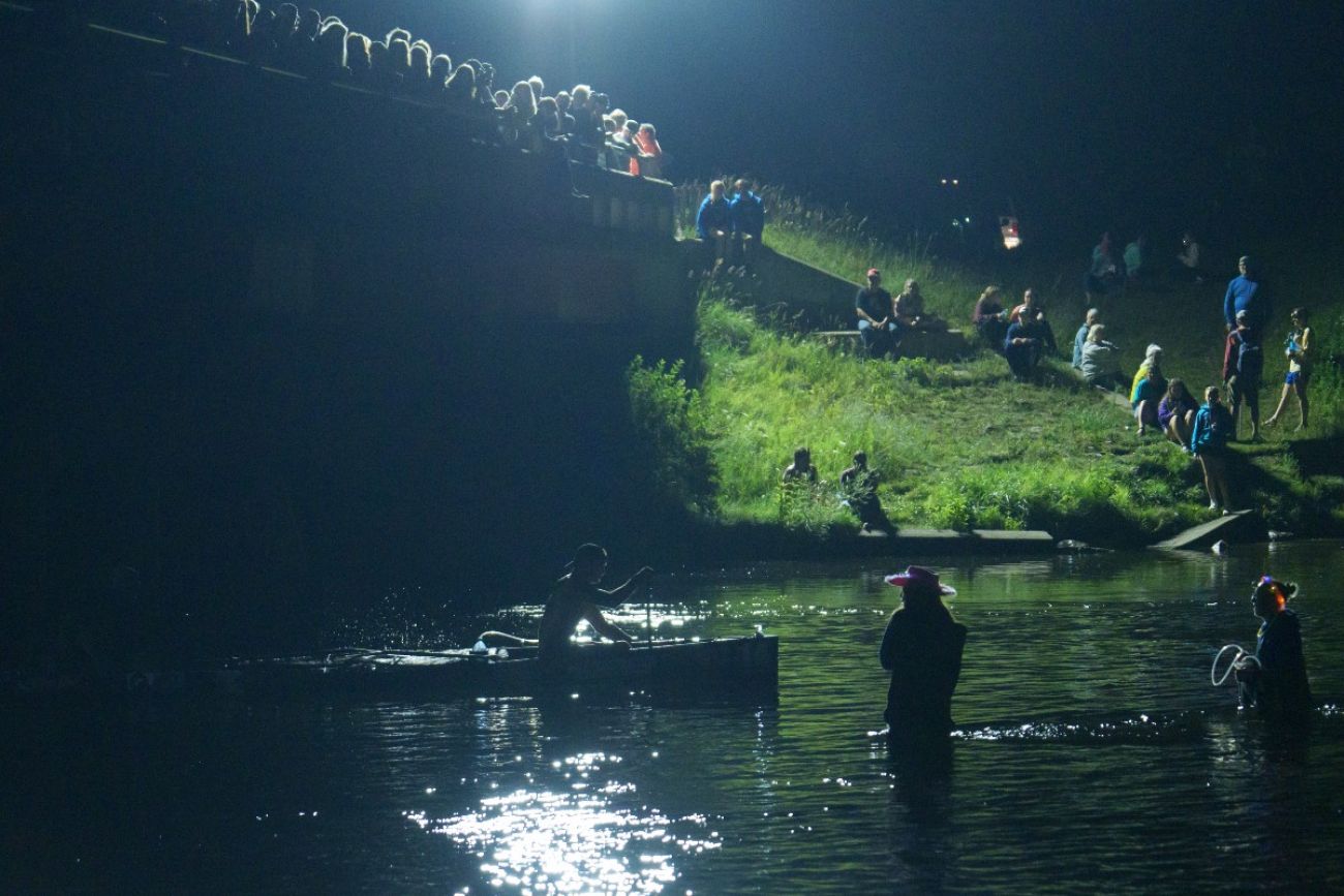 At night, a crowd gathers on a bridge and on a riverbank to watch canoe racers paddle past
