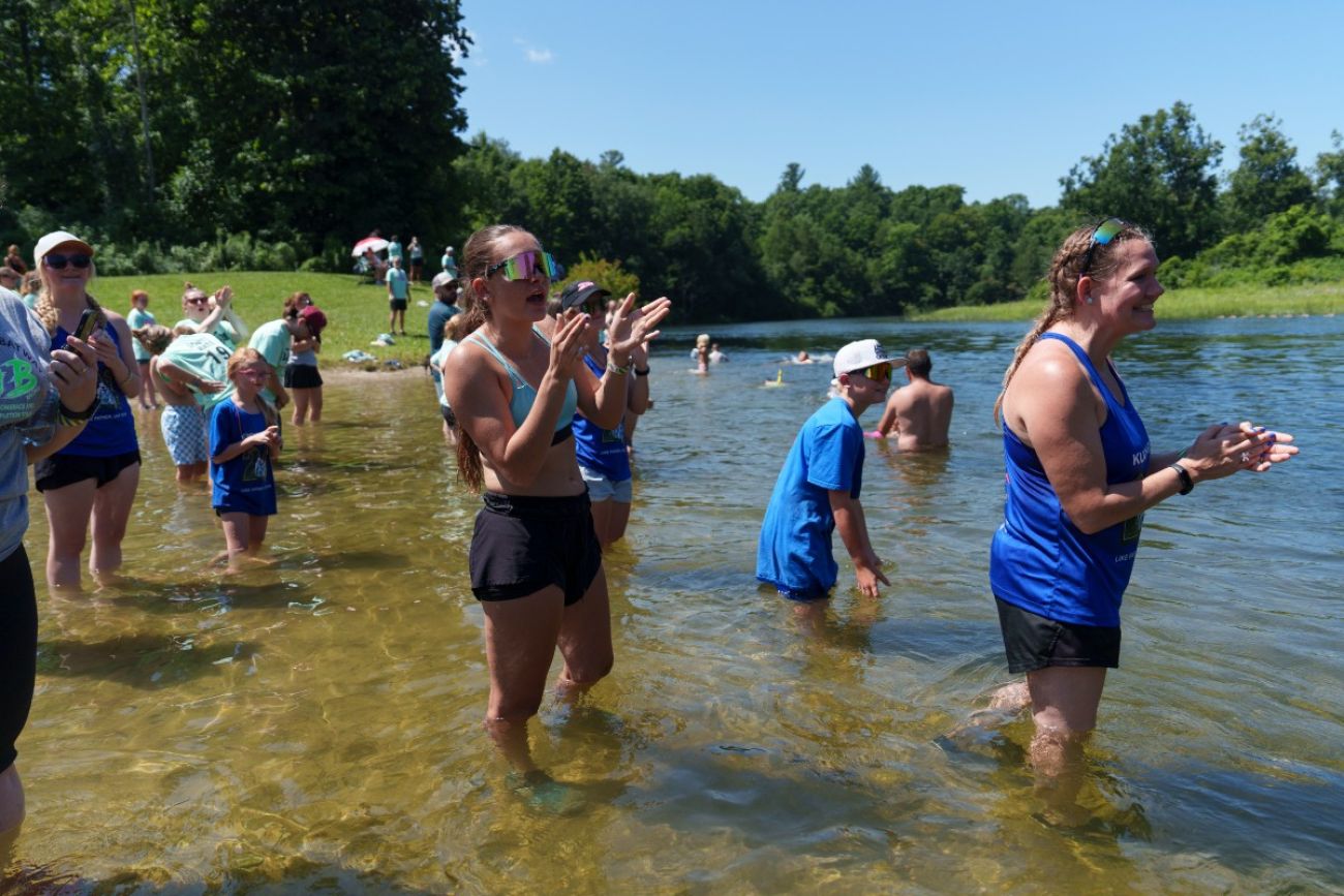 A crowd of people stand shin-deep in a river to cheer on passing canoe racers
