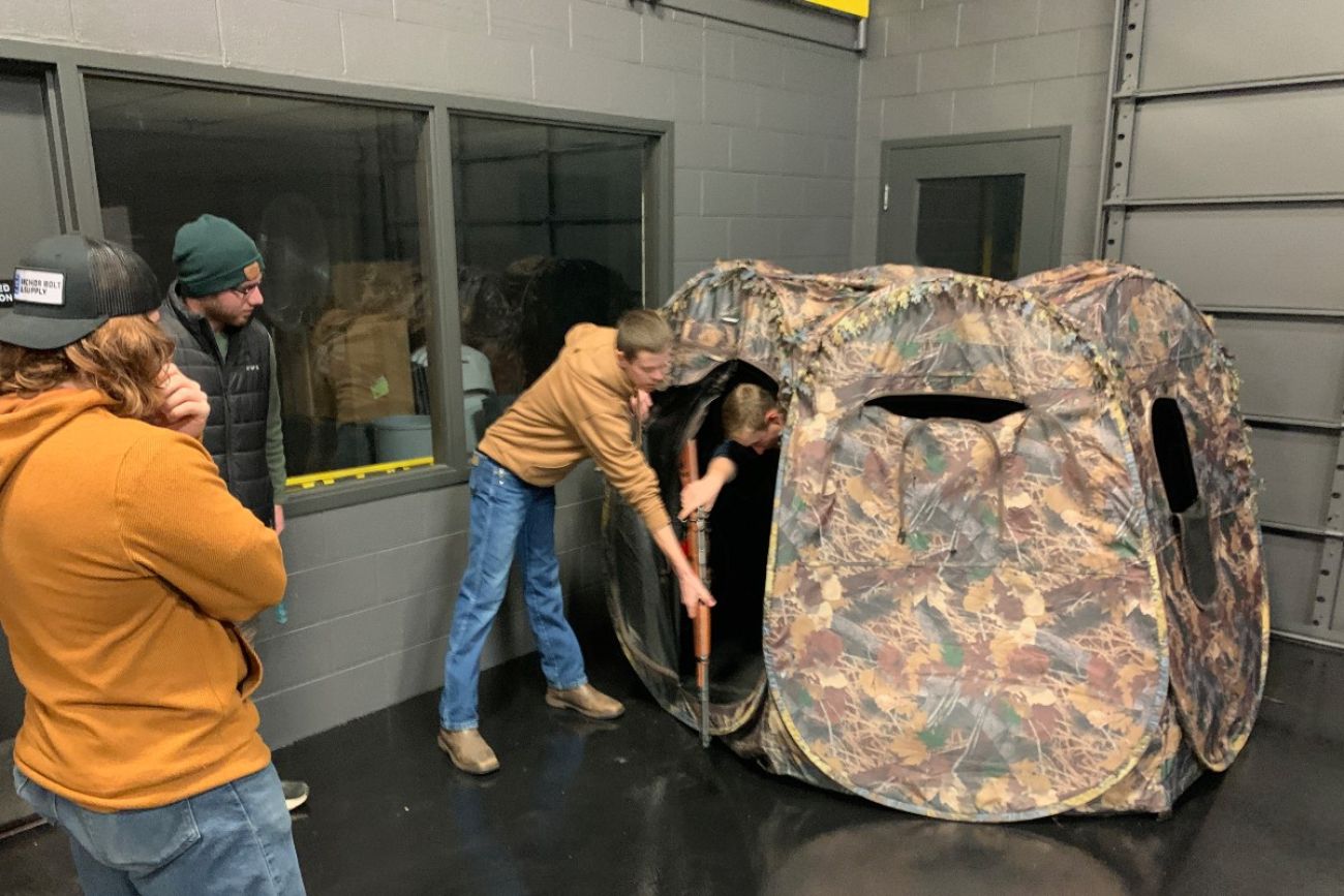 College students stand around a deer blind while another student steps out of the blind holding a rifle
