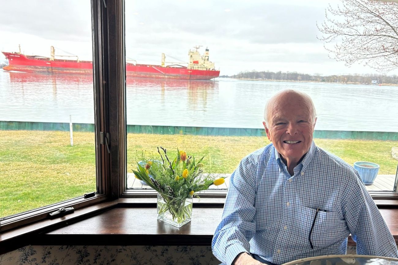A man poses for a photo in front of a window through which a freighter can be seen