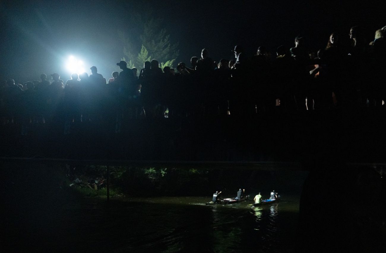 A nighttime scene shows spectators watching canoe racers paddle past
