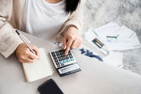  woman writing a list of debt on notebook calculating her expenses with calculator with many invoices 