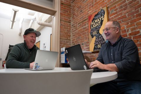 Two men laugh in an office on their computers