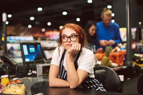 Young boring cashier in eyeglasses and striped apron dreamily looking aside working in modern supermarket with customers on background