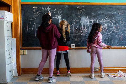 three kids in front of a blackboard. one of the kids is completing a math problem 