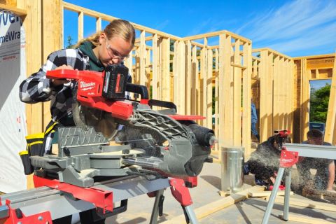 A high school-aged girl works a saw in the foreground while others work in the background of a home under construction