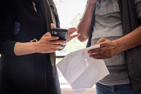 A woman holding a phone talks to a person holding a paper. The photo doesn't show their faces.