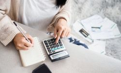  woman writing a list of debt on notebook calculating her expenses with calculator with many invoices 