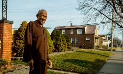 A man wearing brown, standing in front of his house