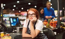 Young boring cashier in eyeglasses and striped apron dreamily looking aside working in modern supermarket with customers on background