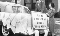 A black and white photo. There's a sign on the back of a car that says, "Stop me if you have not signed a con-con petition." 