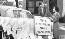 A black and white photo. There's a sign on the back of a car that says, "Stop me if you have not signed a con-con petition." 