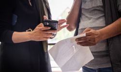 A woman holding a phone talks to a person holding a paper. The photo doesn't show their faces.