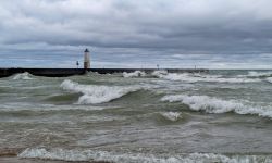 A lake is seen with big waves flowing toward shore and a lighthouse at the end of a pier in the background