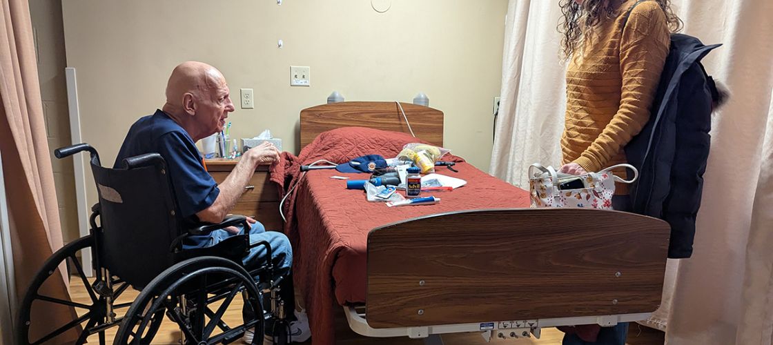 Howard Gregor in a wheelchair by a medical bed. A woman is standing on the other side of the bed.