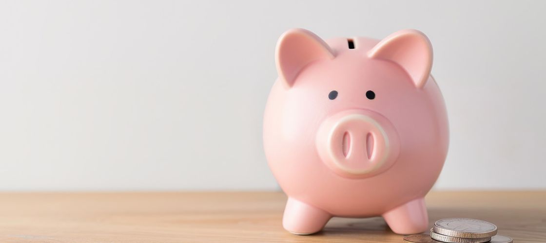A pink piggy bank overflowing with coins is placed on a wooden table, against a white wall background.
