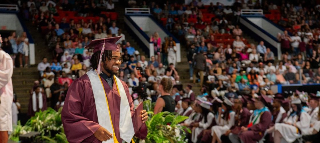 A young man in cap and gown picks up his high school diploma in an auditorium