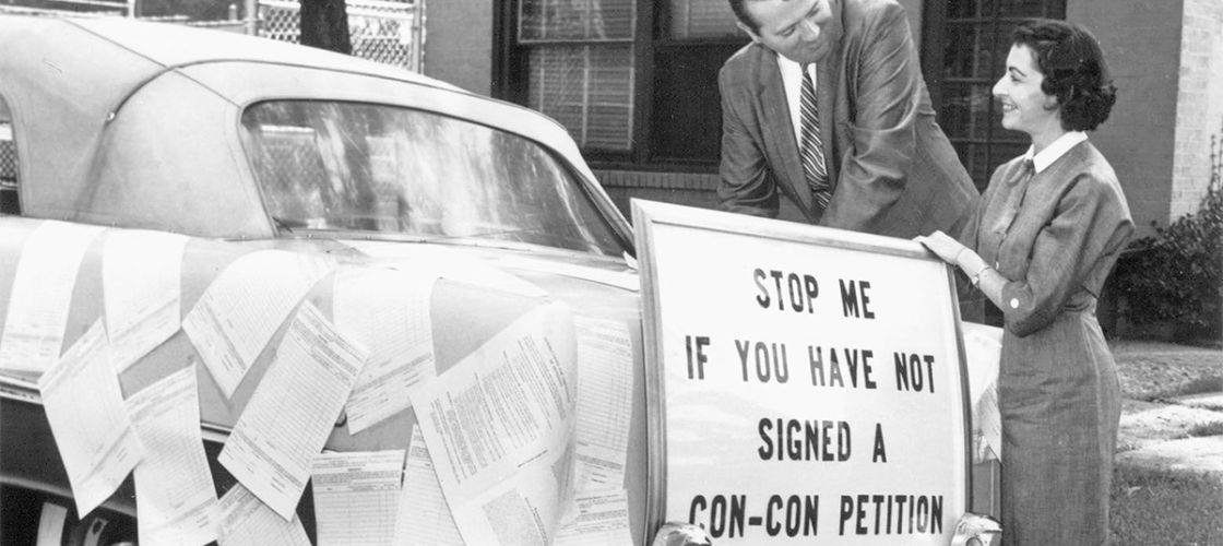 A black and white photo. There's a sign on the back of a car that says, "Stop me if you have not signed a con-con petition." 